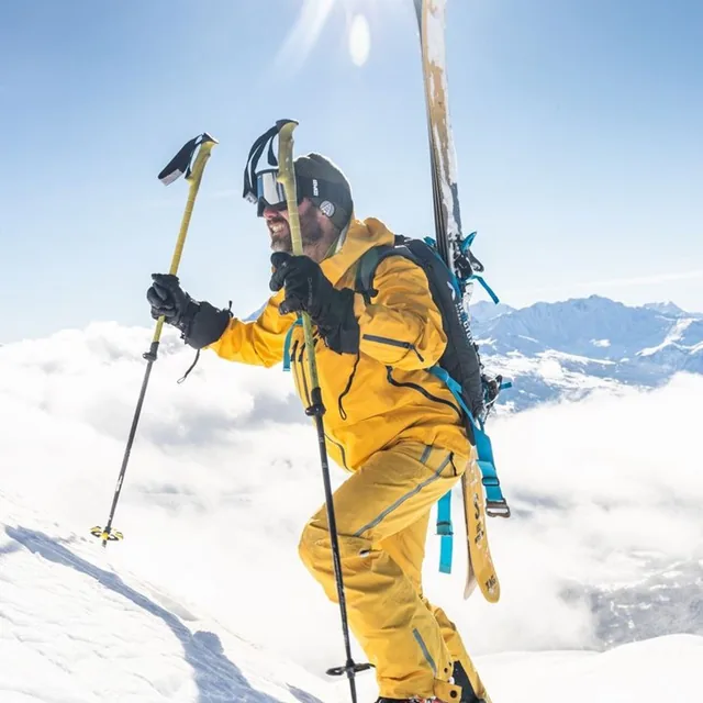 A skier in a yellow suit climbing a snow slope with skis on the pack above a sea of cloud