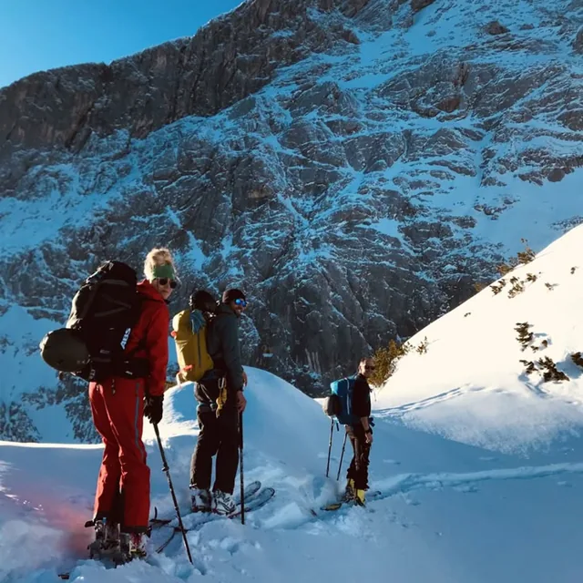 Three ski tourers pausing on a snow slope beneath a steep snow-streaked rock face