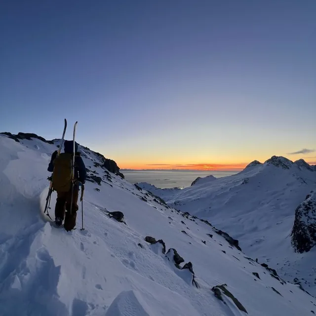 A ski tourer carrying skis on their pack climbing a snow ridge at dawn with the sun low on the horizon