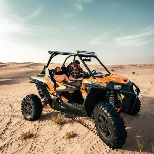 An orange and black two-seater buggy with helmeted riders parked on soft desert sand
