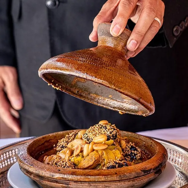 A chef lifting a clay tagine lid to reveal chicken with almonds, sesame and preserved lemon