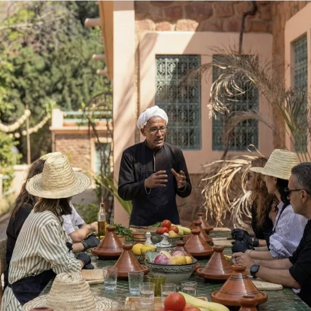 A chef in a white turban and apron explaining the recipe to guests in straw hats seated around a tagine-covered table