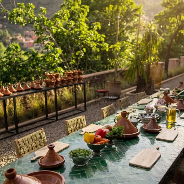 A long green tiled cooking table set with clay tagines, chopping boards and vegetables in a garden below the mountains