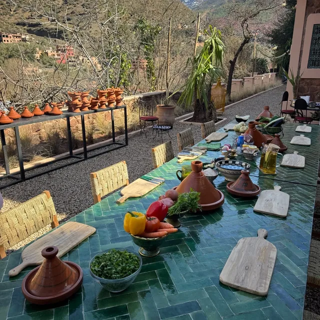 A long green tiled cooking table laid with clay tagines, boards and vegetables on a terrace overlooking a mountain valley village