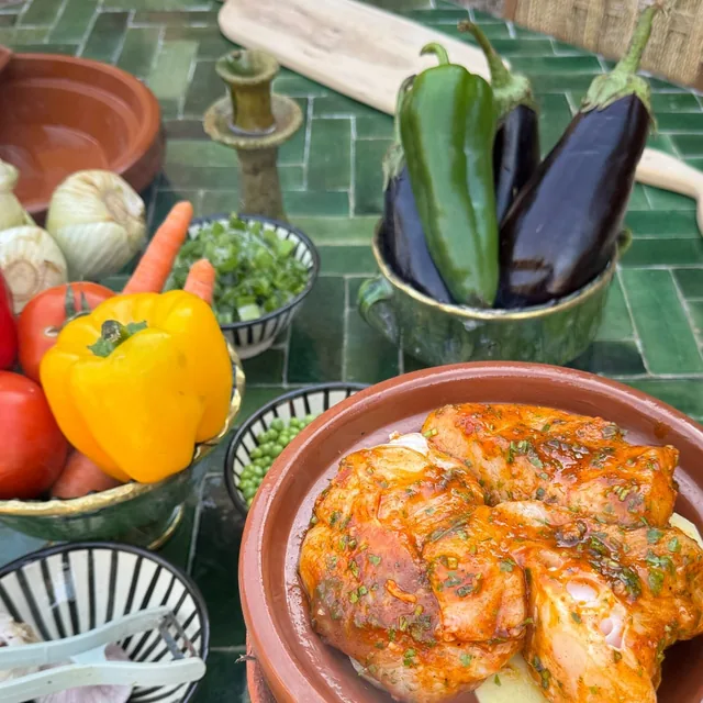 Marinated chicken in a clay tagine dish beside bowls of peppers, aubergines, tomatoes and herbs on a green tiled table