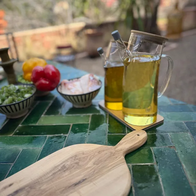 Glass jugs of olive oil beside an olive-wood board and bowls of chopped vegetables on a green tiled table
