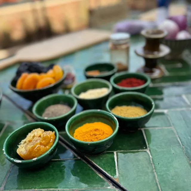 Small green ceramic bowls of turmeric, paprika, cumin, ginger, preserved lemon and dried fruit on a tiled table