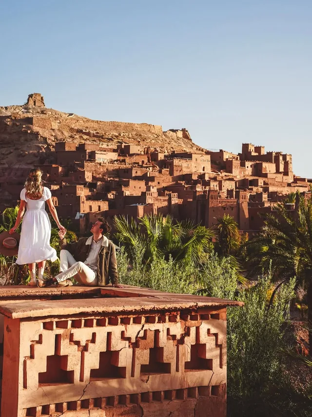 Two travellers resting on a kasbah terrace facing the tiered earth houses of Aït Ben Haddou