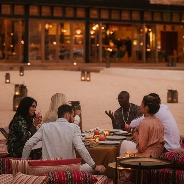 Friends sharing dinner on low sofas at a lantern-lit desert camp at dusk