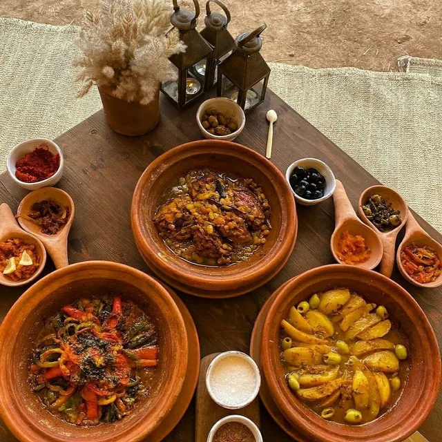 Three clay tagines with vegetables, chicken and potatoes surrounded by small salad bowls and lanterns on a wooden desert table