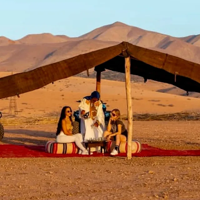 Travellers sitting on rugs and cushions under a black nomad tent taking mint tea below desert dunes