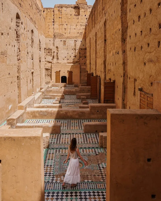 A visitor walking the zellige mosaic floors between the high earth walls of El Badi Palace