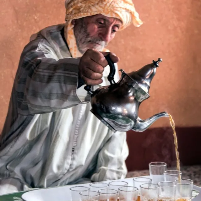 An elderly man in a striped djellaba pouring mint tea from a silver pot into a tray of glasses