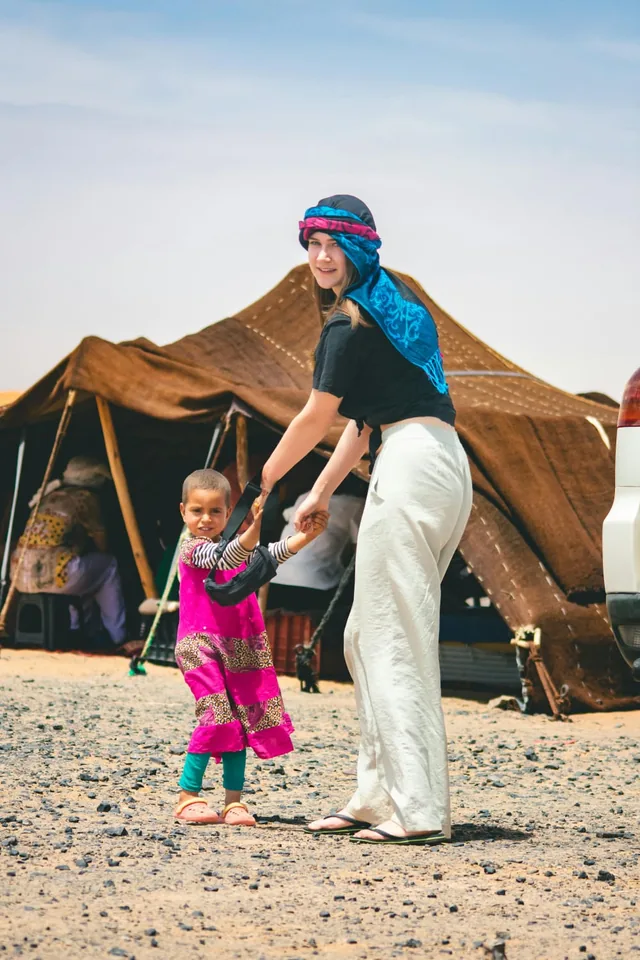 A guest in a blue desert turban holding hands with a local child outside a brown nomad tent