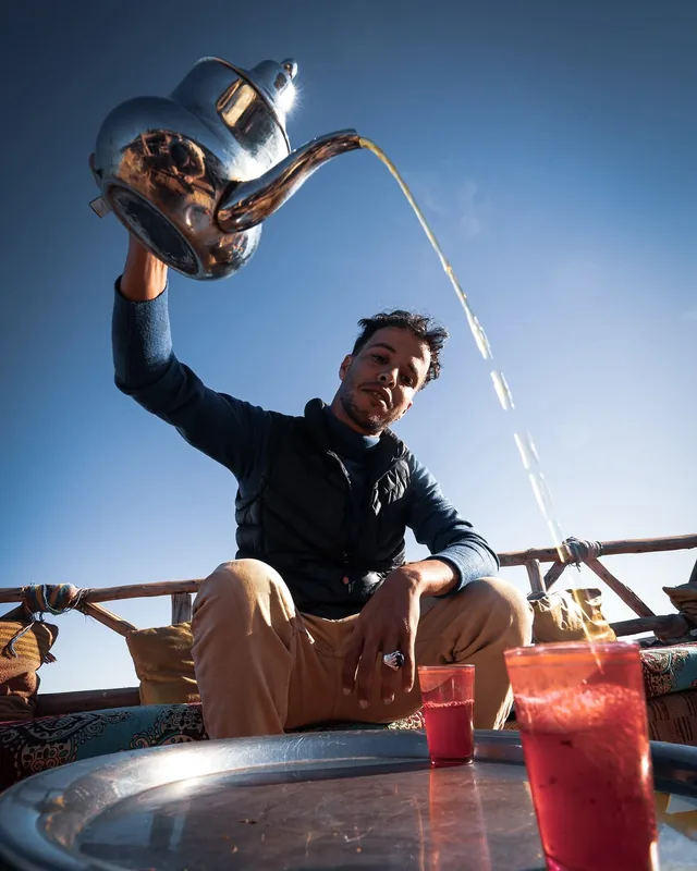 A host pouring mint tea from a raised silver pot into glasses on a sunlit terrace