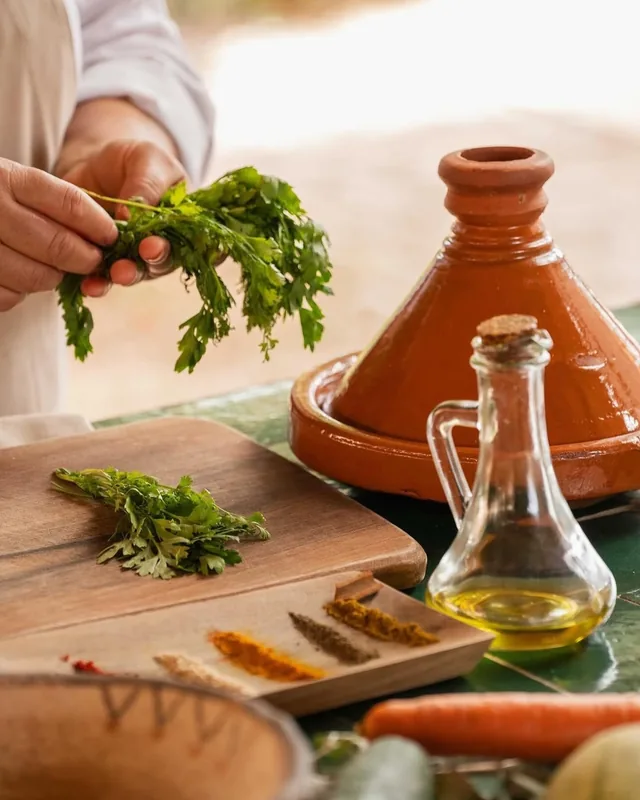 Hands holding fresh parsley beside a clay tagine, olive oil and lines of ground spices