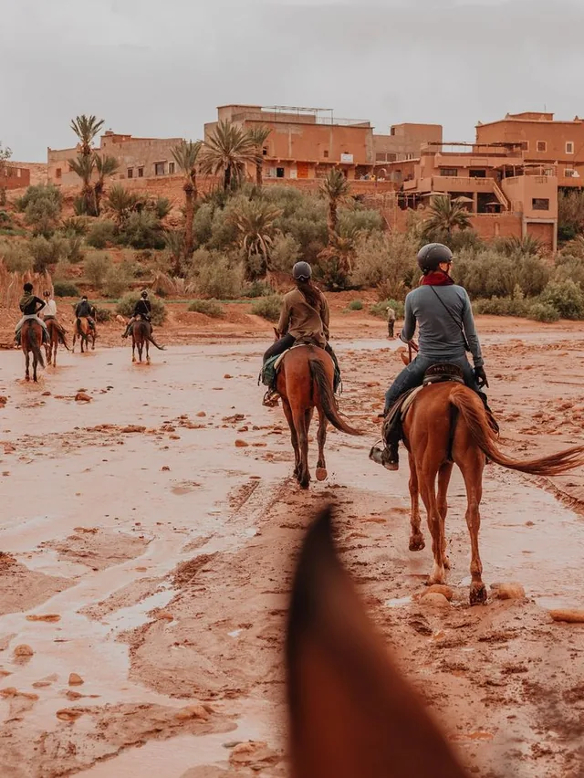 Horse riders crossing a wide red riverbed below an earthen village with palm trees