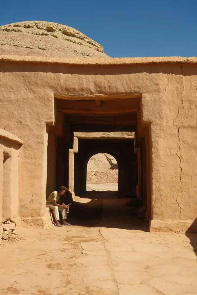 Man resting in the shade of an earthen kasbah passage under a bright blue sky