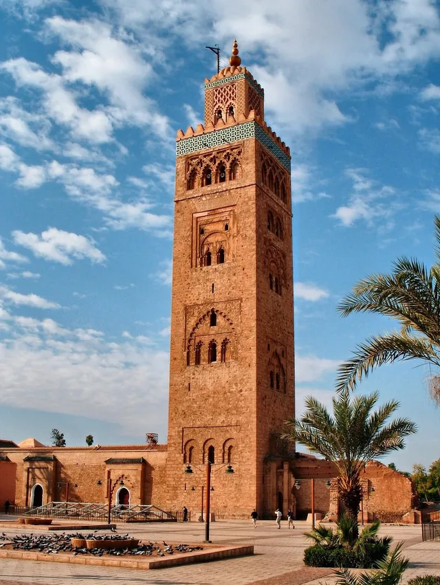 The Koutoubia mosque minaret in warm stone with palm trees under a bright cloudy sky