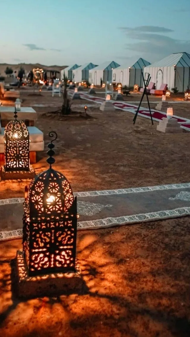 Lit metal lanterns and rugs in front of white desert camp tents at dusk