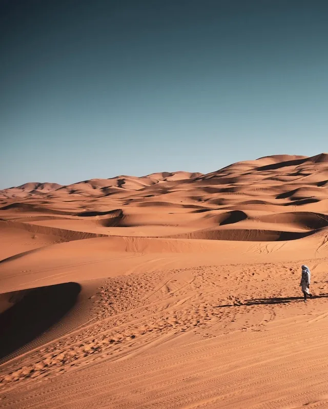 A lone figure in white robes crossing a vast expanse of Sahara dunes under a deep blue sky