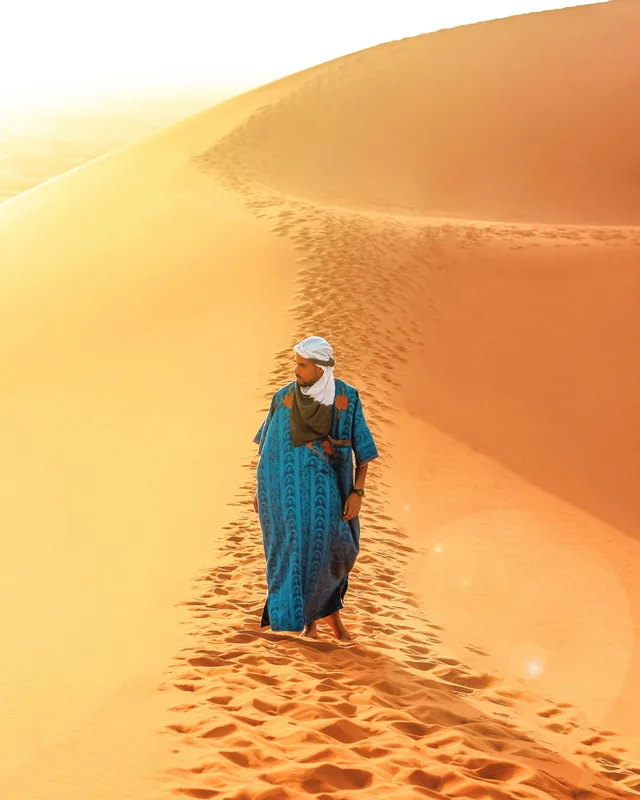 A man in a blue robe and turban walking a footprint-marked dune ridge at sunrise