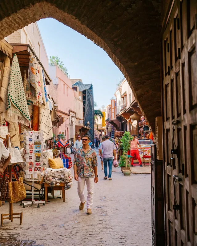 View through a stone arch onto a busy Marrakech medina street lined with rug and craft stalls