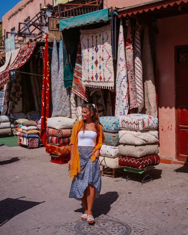 Traveller walking past hanging Berber rugs and stacked floor cushions in a Marrakech rug souk