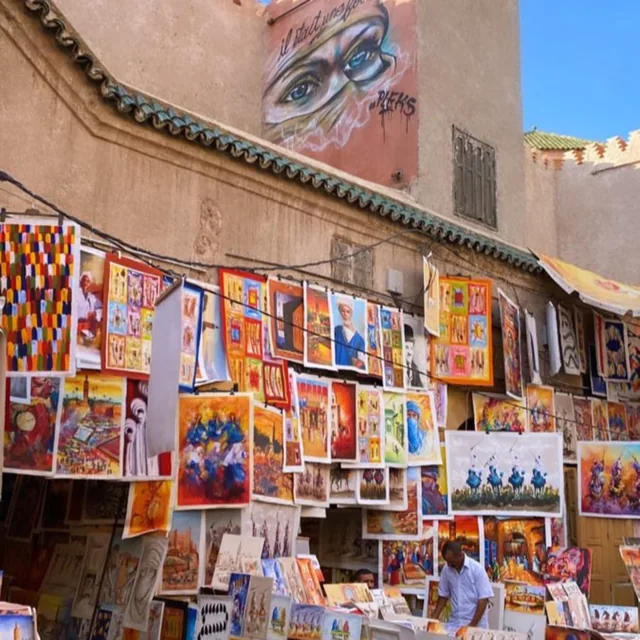 Colourful paintings hung along a sunlit medina wall below a large street-art mural