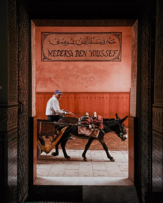A man driving a donkey cart past the Medersa Ben Youssef sign in the Marrakech medina