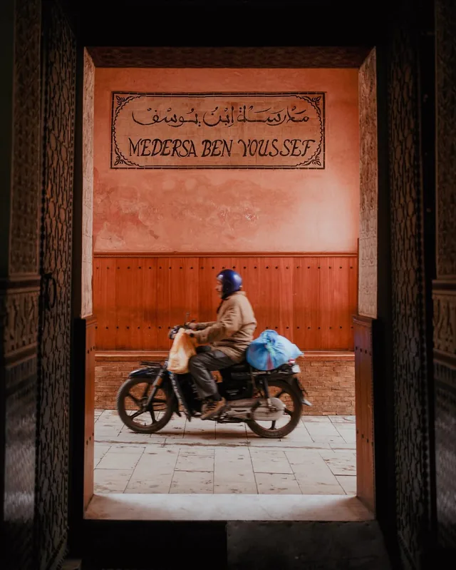 A motorbike passing the tiled Medersa Ben Youssef sign, seen through a carved medina doorway