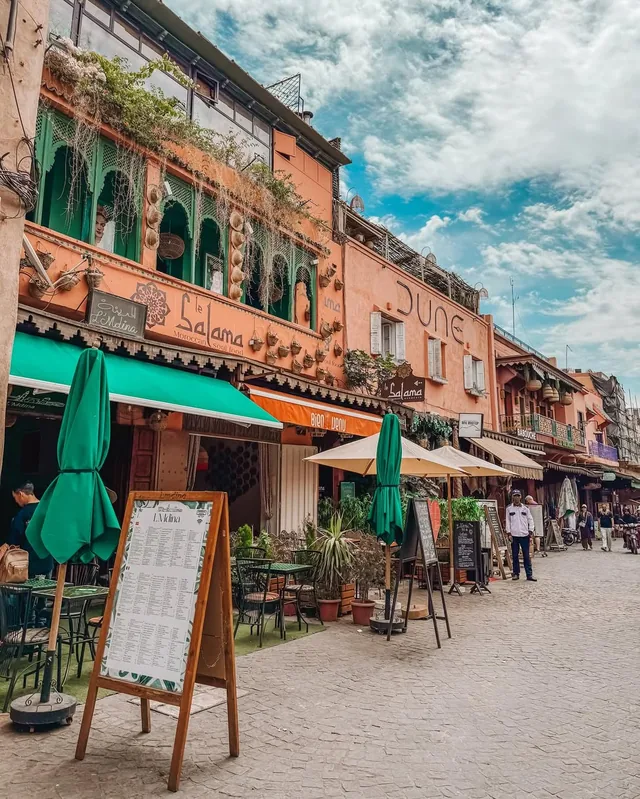 Café terraces with parasols along a pink Marrakech medina street under a bright sky