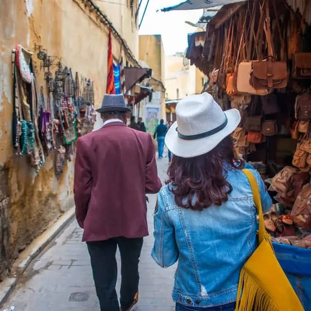 Two people walking a narrow medina lane past hanging leather bags and handmade jewellery