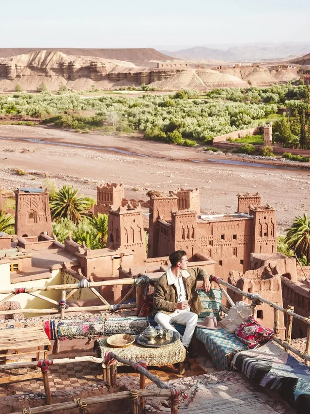 A traveller with a silver mint tea tray on a cushioned terrace above the kasbahs of Aït Ben Haddou