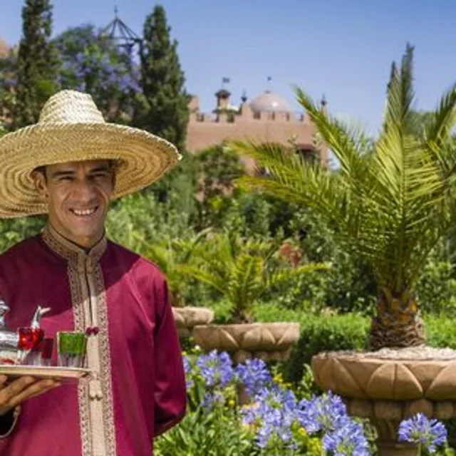 A smiling host in a straw hat carrying a tray of mint tea through a Marrakech garden