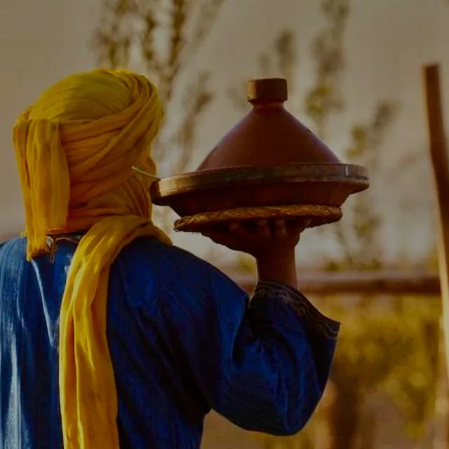 A host in a blue robe and yellow turban carrying a covered clay tagine to the table