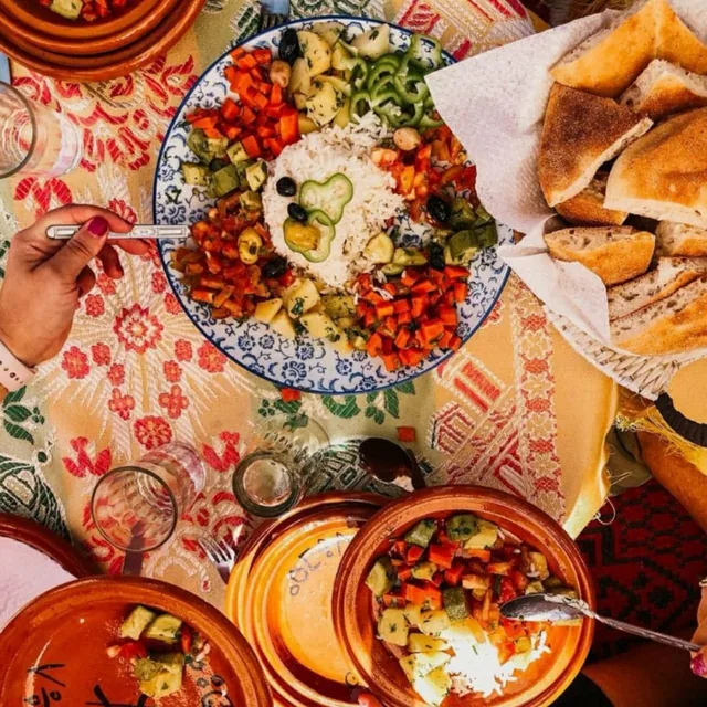 A shared Moroccan lunch table with a rice and vegetable platter, tagines and fresh khobz bread on an embroidered cloth