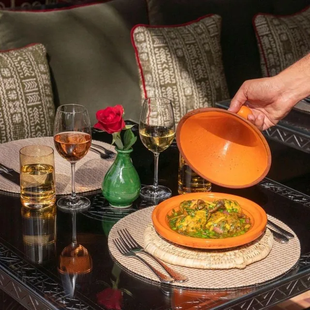 A server lifting the lid of a tagine on a low table set with glasses of wine and a red rose