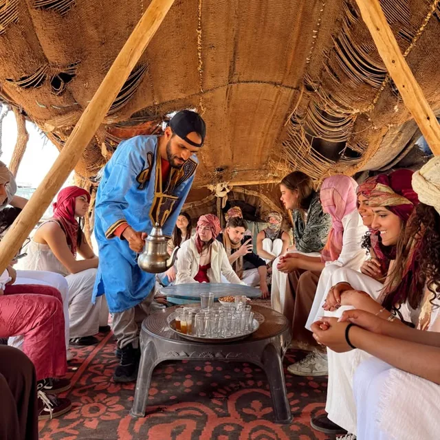 A host in a blue tunic pouring mint tea for a group of travellers seated on rugs inside a nomad tent