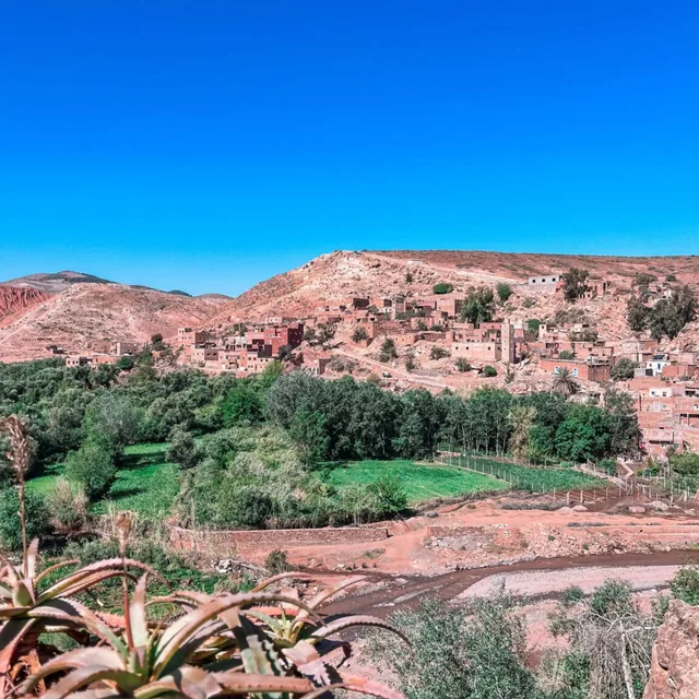 A red earth Berber village built into the hillside above green fields and a river bed in the Atlas foothills
