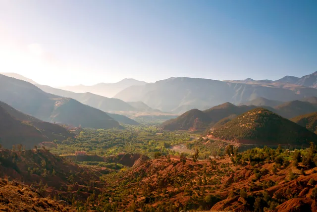 Green valley floor and red earth slopes below the hazy ridges of the High Atlas