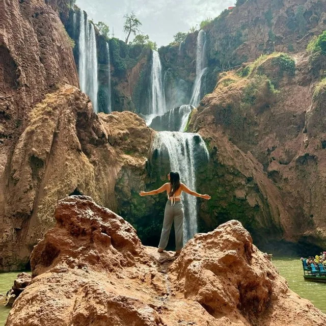A traveller standing on red rocks with arms open in front of the multi-tier Ouzoud waterfalls