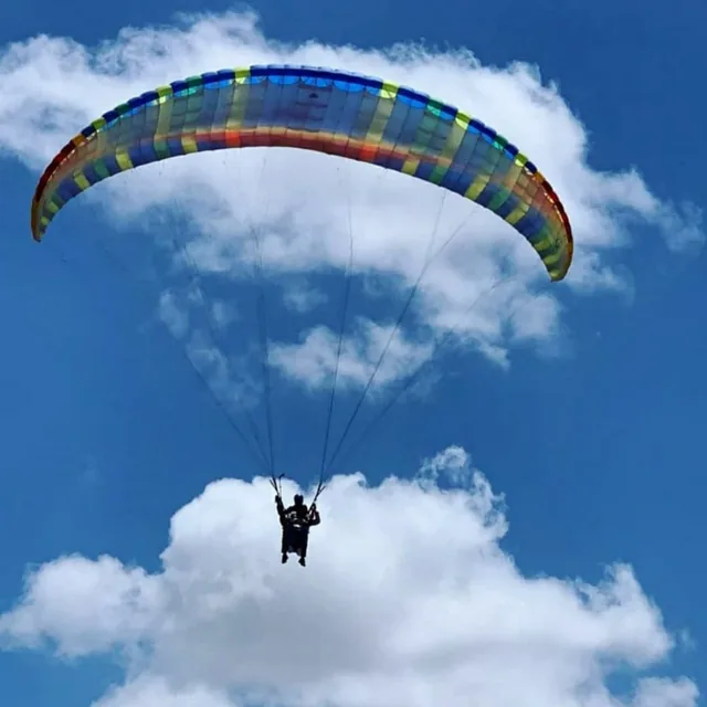 A tandem paraglider flying under a rainbow-coloured wing among white clouds in a deep blue sky