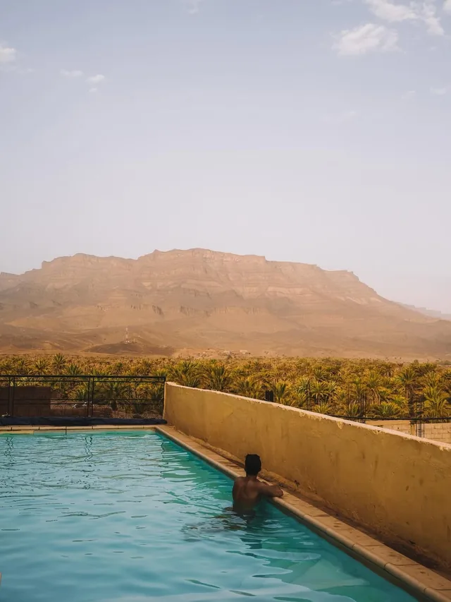 A guest in a long pool above a date palm grove with the Draa valley cliffs beyond