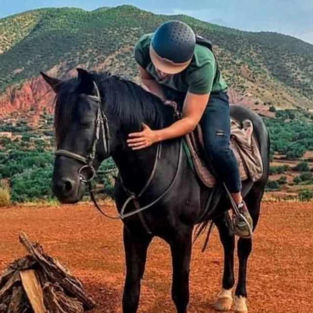 A rider in a helmet leaning down to stroke a black horse in front of green Atlas foothills