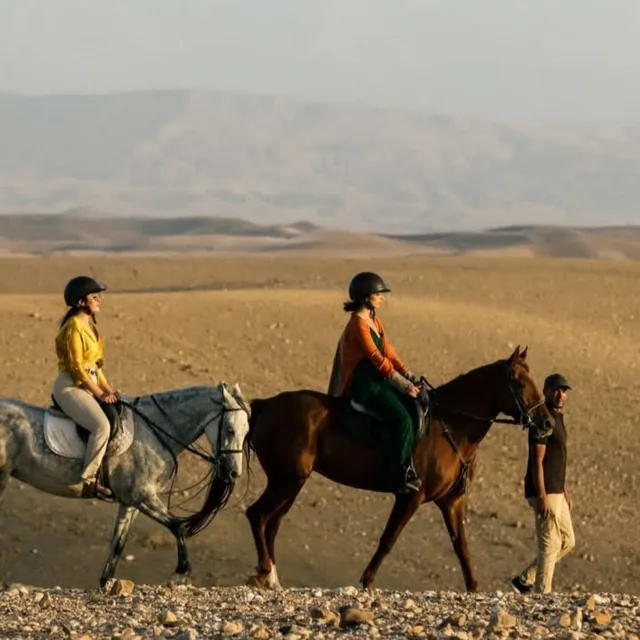 Two riders on grey and chestnut horses walking the open Agafay plain beside their guide