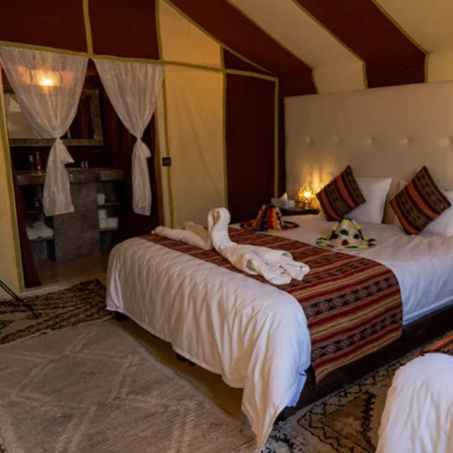 A striped nomad tent with twin beds, Berber rugs and a lit lantern beside the bathroom