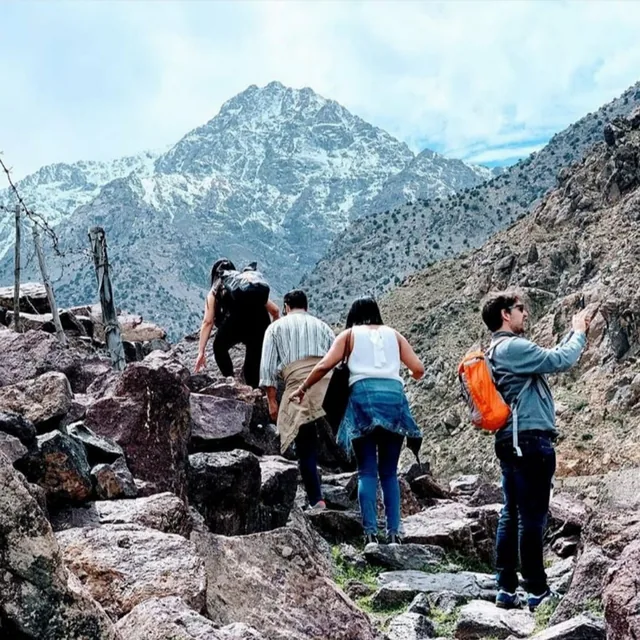 A small group climbing rough stone steps on a High Atlas trail with the snow-covered Toubkal massif ahead