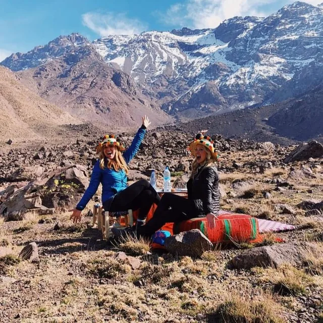 Two trekkers in Berber straw hats sitting at a rug picnic on the mountainside below snow-covered Atlas peaks