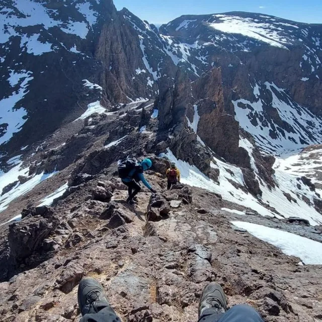 Two trekkers scrambling along a rocky Toubkal ridge with snow patches on the slopes below